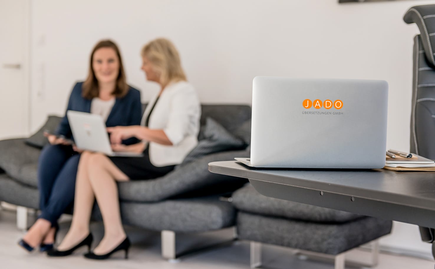 Dieses professionelle Businessfoto von Mica Zeitz zeigt zwei Frauen bei einem Meeting, die an einem Sofa sitzen und an Laptops arbeiten, während der JADO Übersetzungen GmbH Laptop im Vordergrund gut sichtbar ist. Ideal für Unternehmen in Ingolstadt und Pfaffenhofen, die hochwertige Businessfotos für ihre Marketingstrategie suchen.