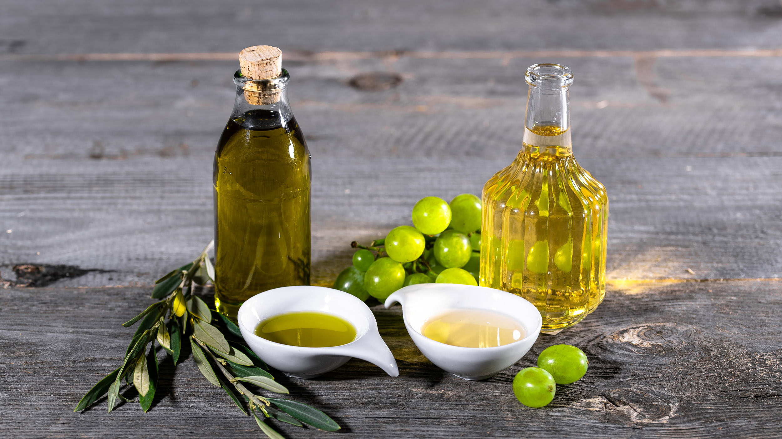 Diese ansprechende Food-Fotografie zeigt mehrere Flaschen mit Olivenöl und eine Schale mit verschiedenen Ölen, umgeben von frischen Trauben. Die rustikale Holzunterlage ergänzt das stilvolle Arrangement und hebt die Qualität der Produkte hervor, ideal für kreative Koch- oder Rezeptideen.