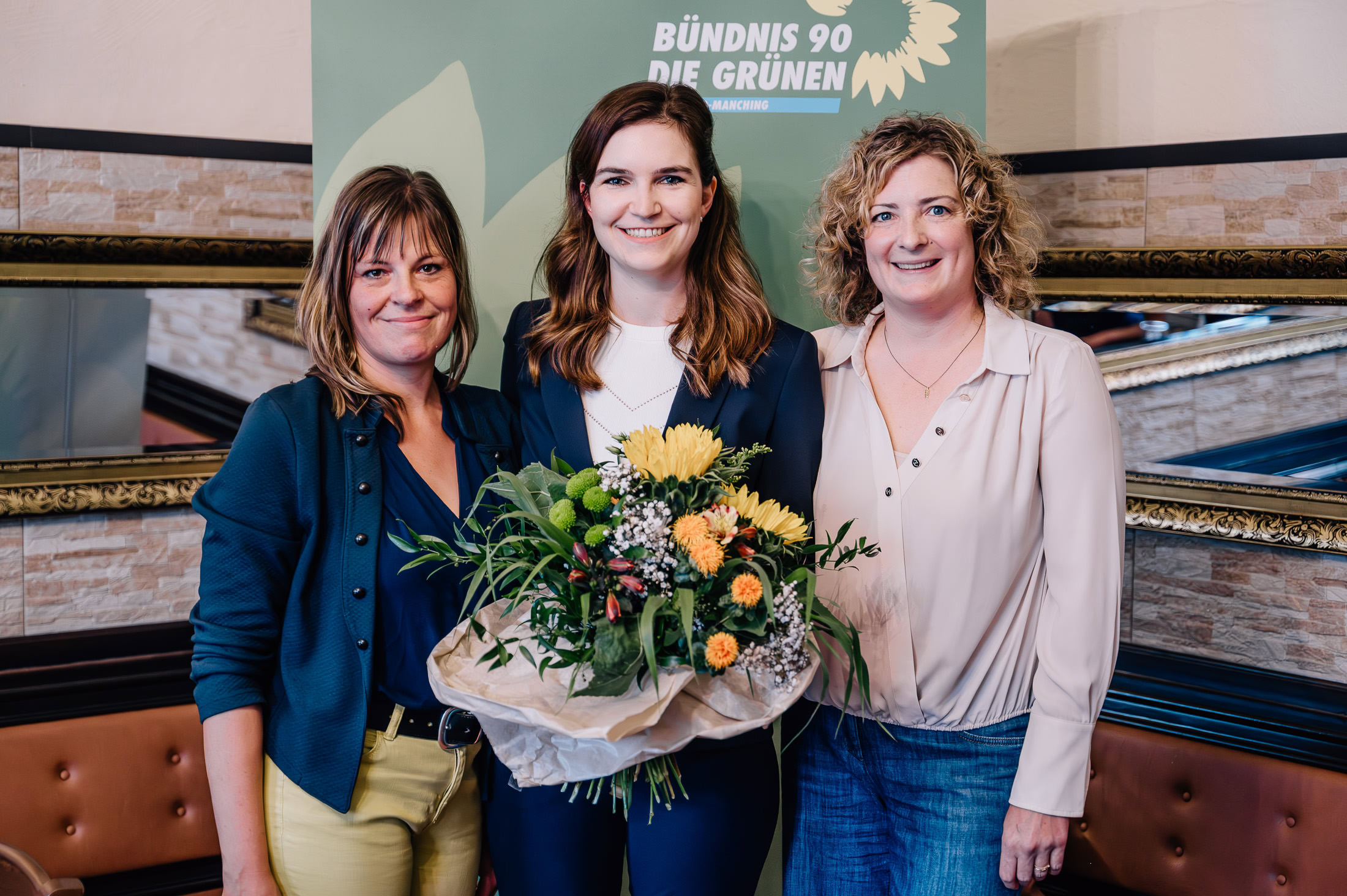 Das Bild zeigt drei Frauen, die lächelnd vor einem Hintergrund mit der Aufschrift "Bündnis 90 Die Grünen" stehen und einen Blumenstrauß halten. Fotografiert von Mica Zeitz, erwecken diese Businessfotos eine positive und einladende Atmosphäre.
